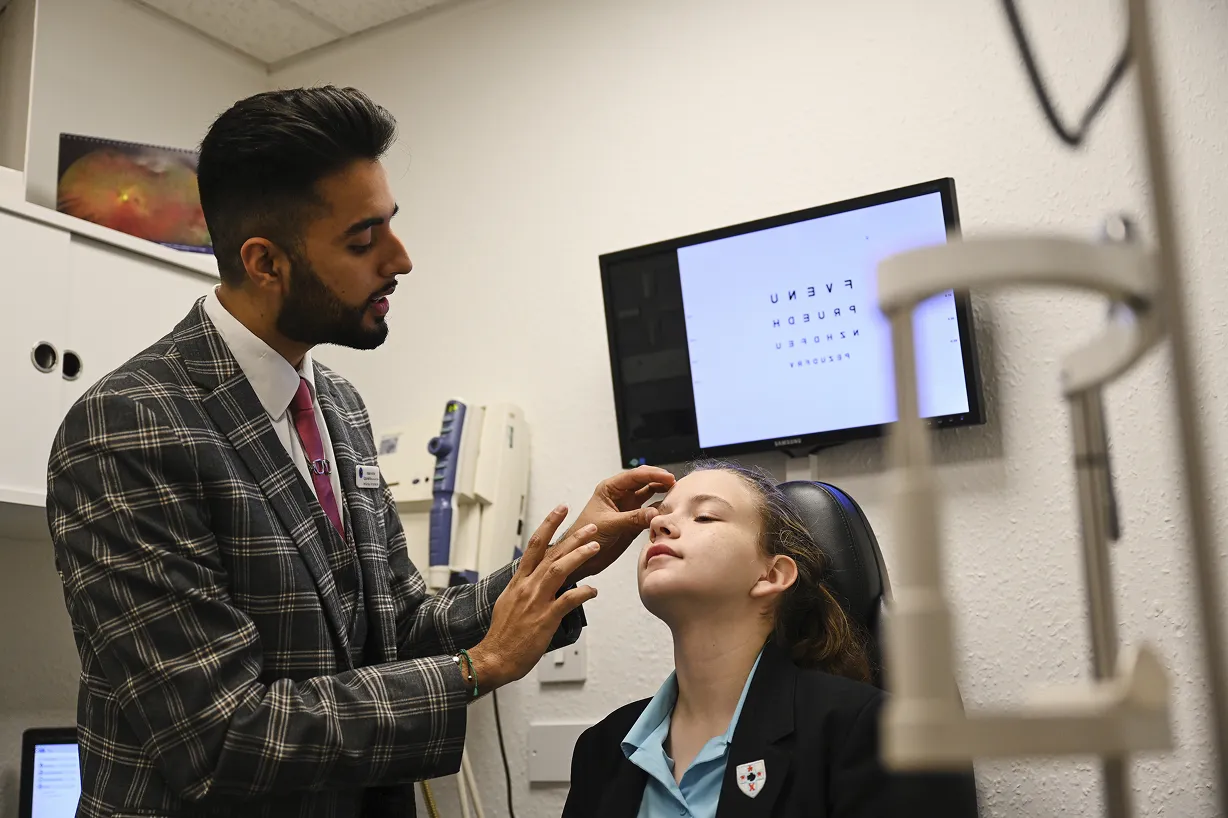 Optometrist examining a young female patient's eyes in an eye clinic with an eye chart displayed on a screen behind them.