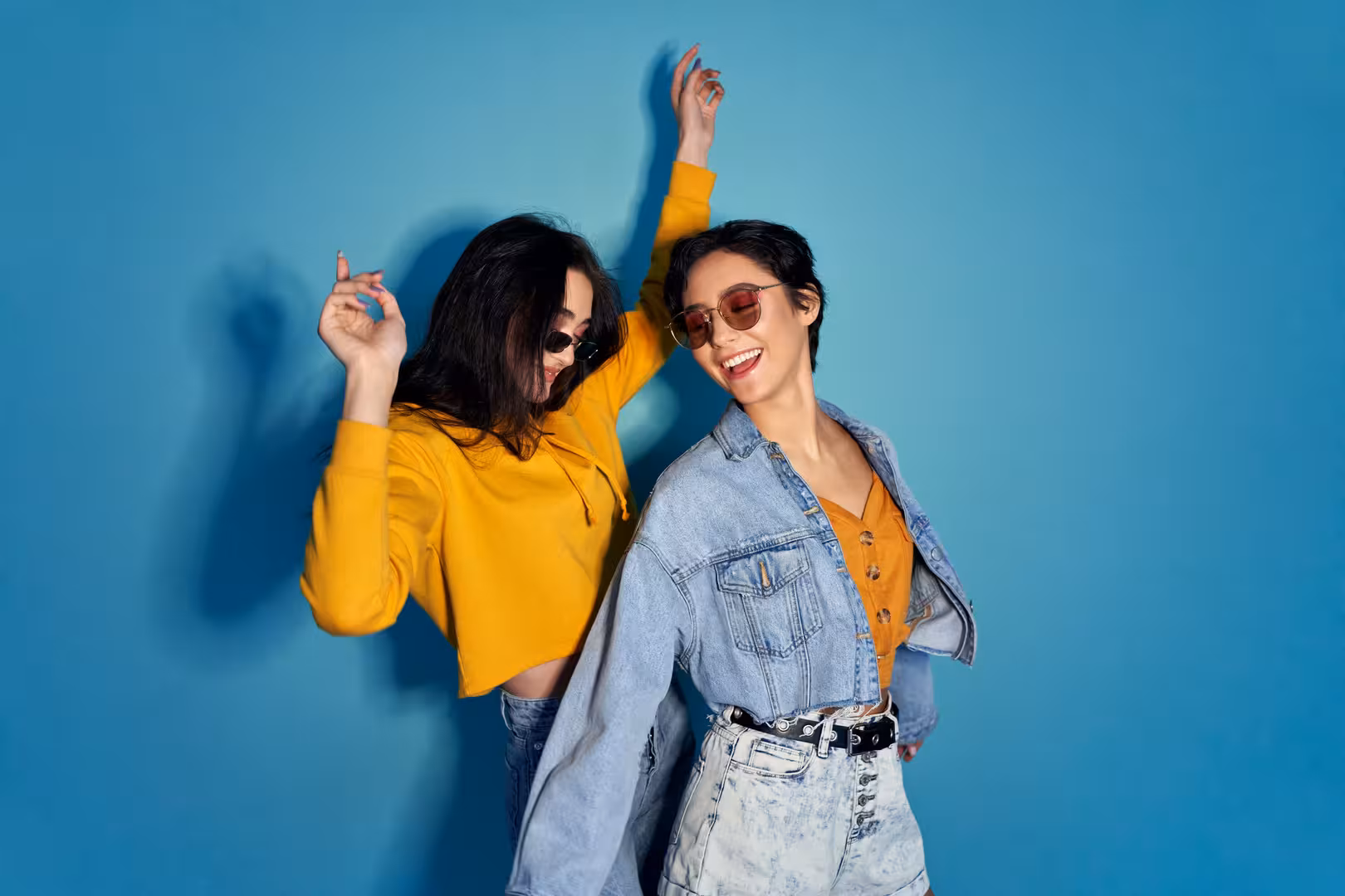 Two women wearing sunglasses and casual outfits dancing against a blue background.