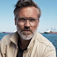 Middle-aged man with glasses and gray hair wearing a beige jacket by the sea with ships in the background.