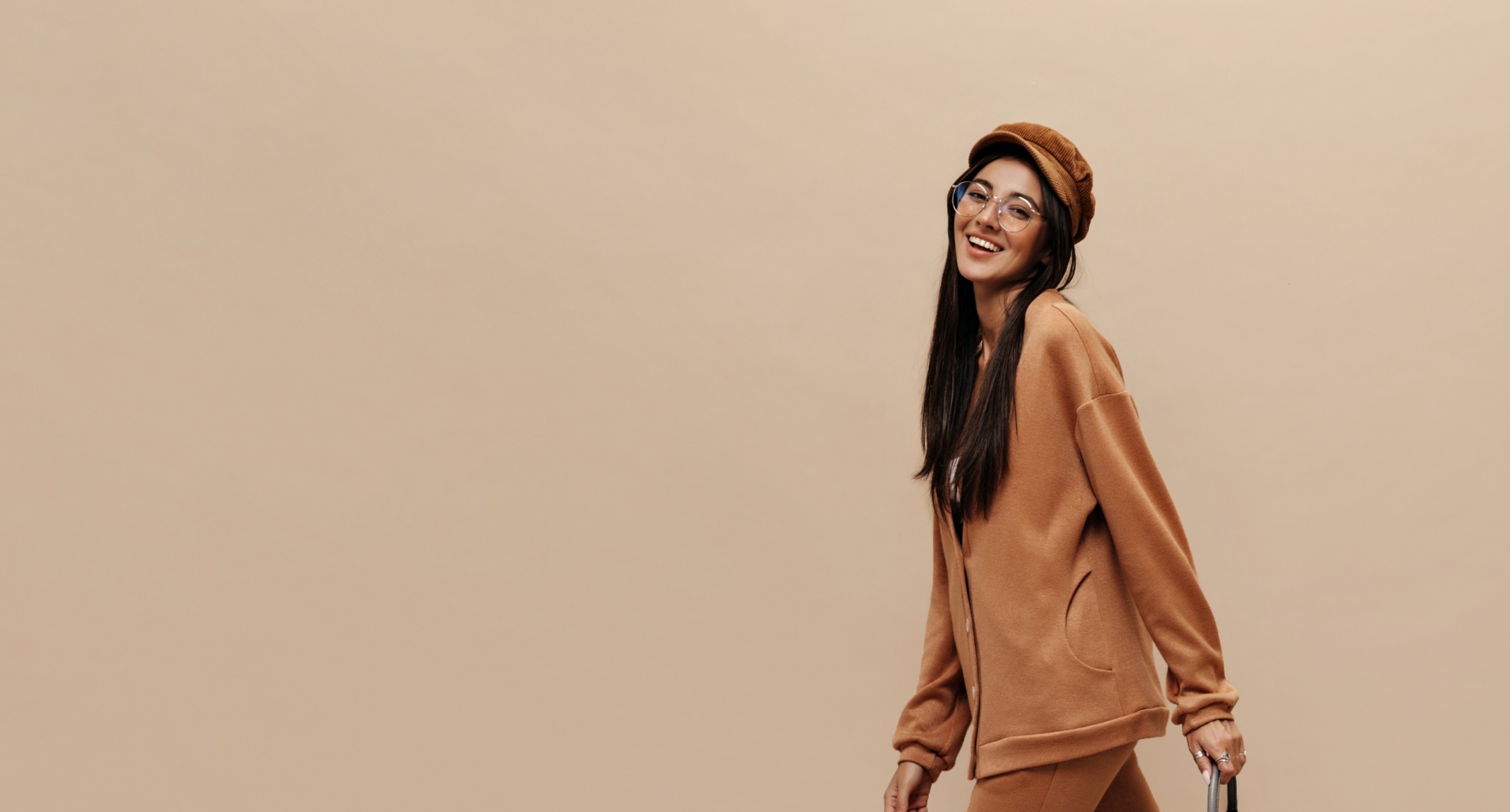 Smiling woman with long dark hair wearing round glasses, a brown hat, and a matching brown outfit, holding a luggage handle.