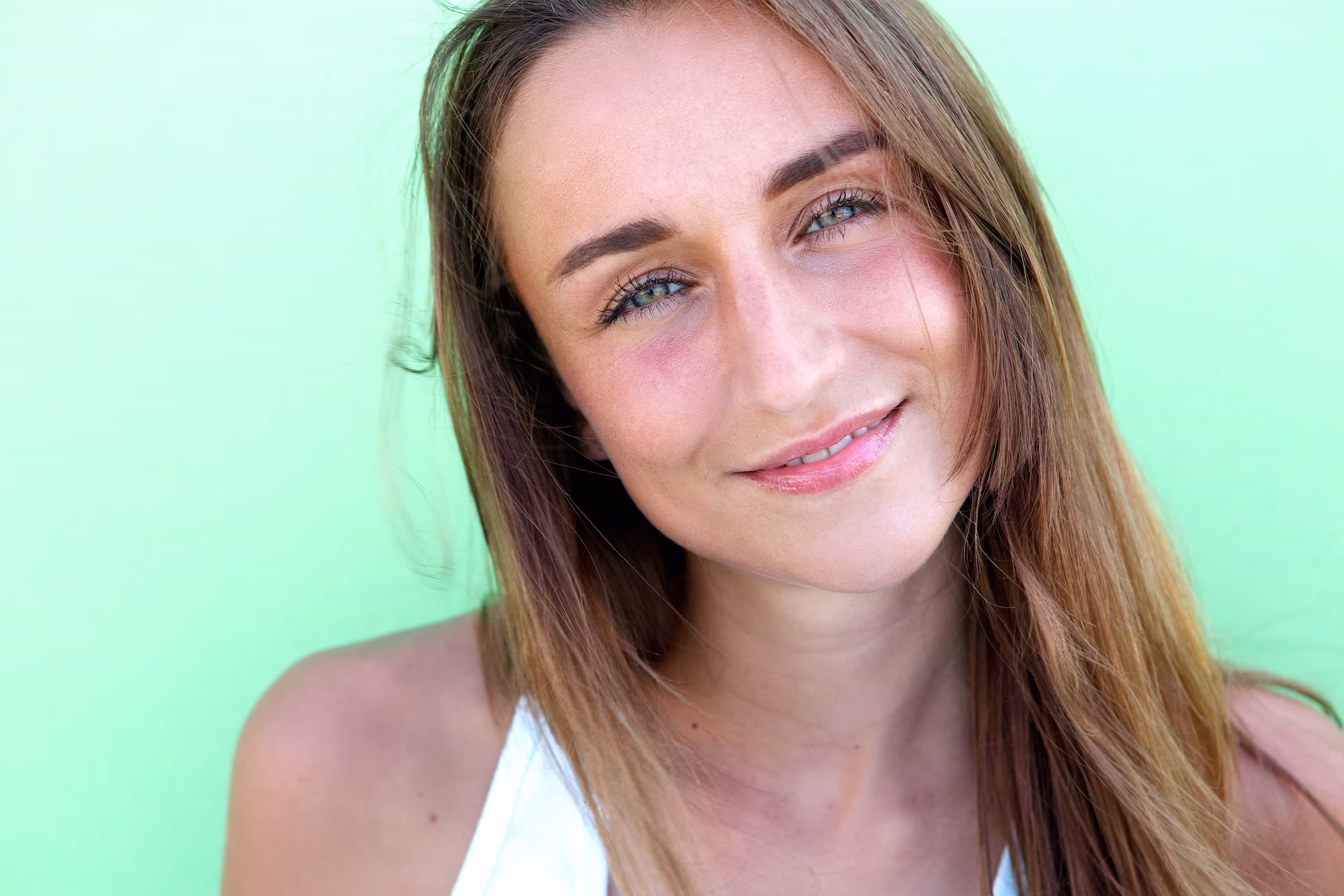 Close-up of a smiling young woman with long brown hair and blue eyes against a green background.