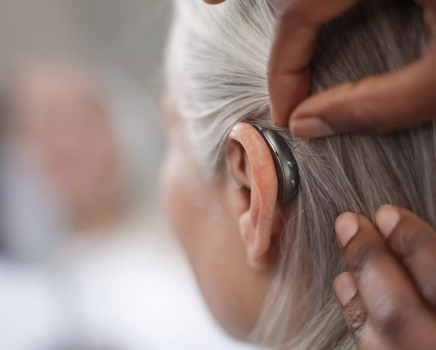 Close-up of hands adjusting a hearing aid on the ear of an elderly person with gray hair.