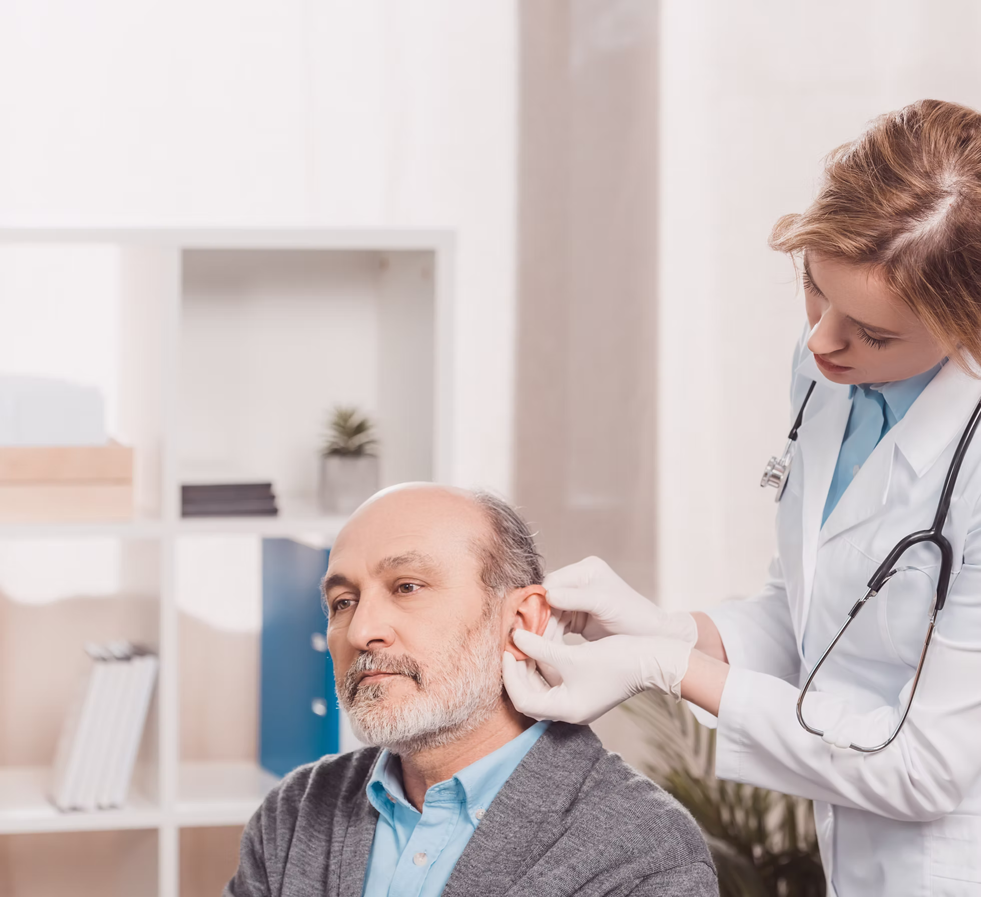 Female doctor wearing gloves adjusts a hearing aid in an older man's ear in a medical office.