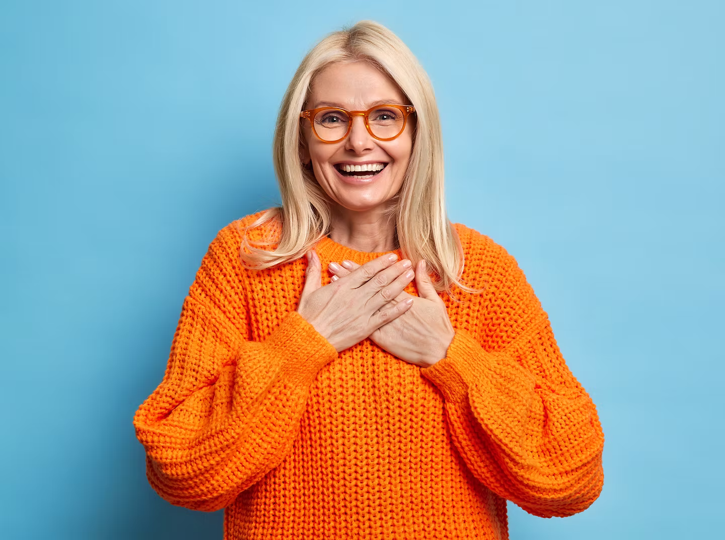 Smiling blond woman wearing orange sweater and glasses, touching her chest with both hands against a blue background.