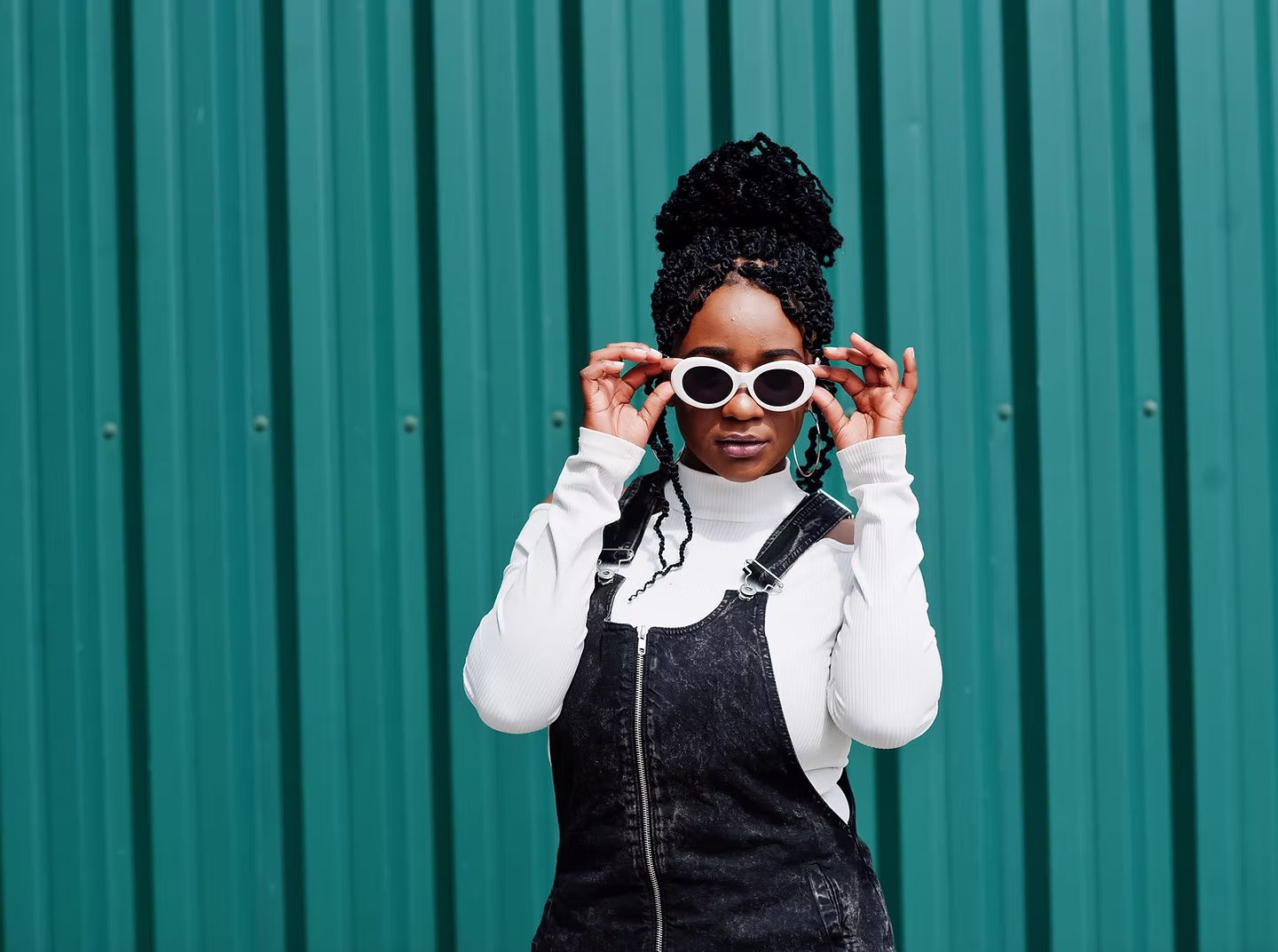 Woman with braided hair wearing white sunglasses, a white turtleneck, and black denim overalls against a teal corrugated metal background.