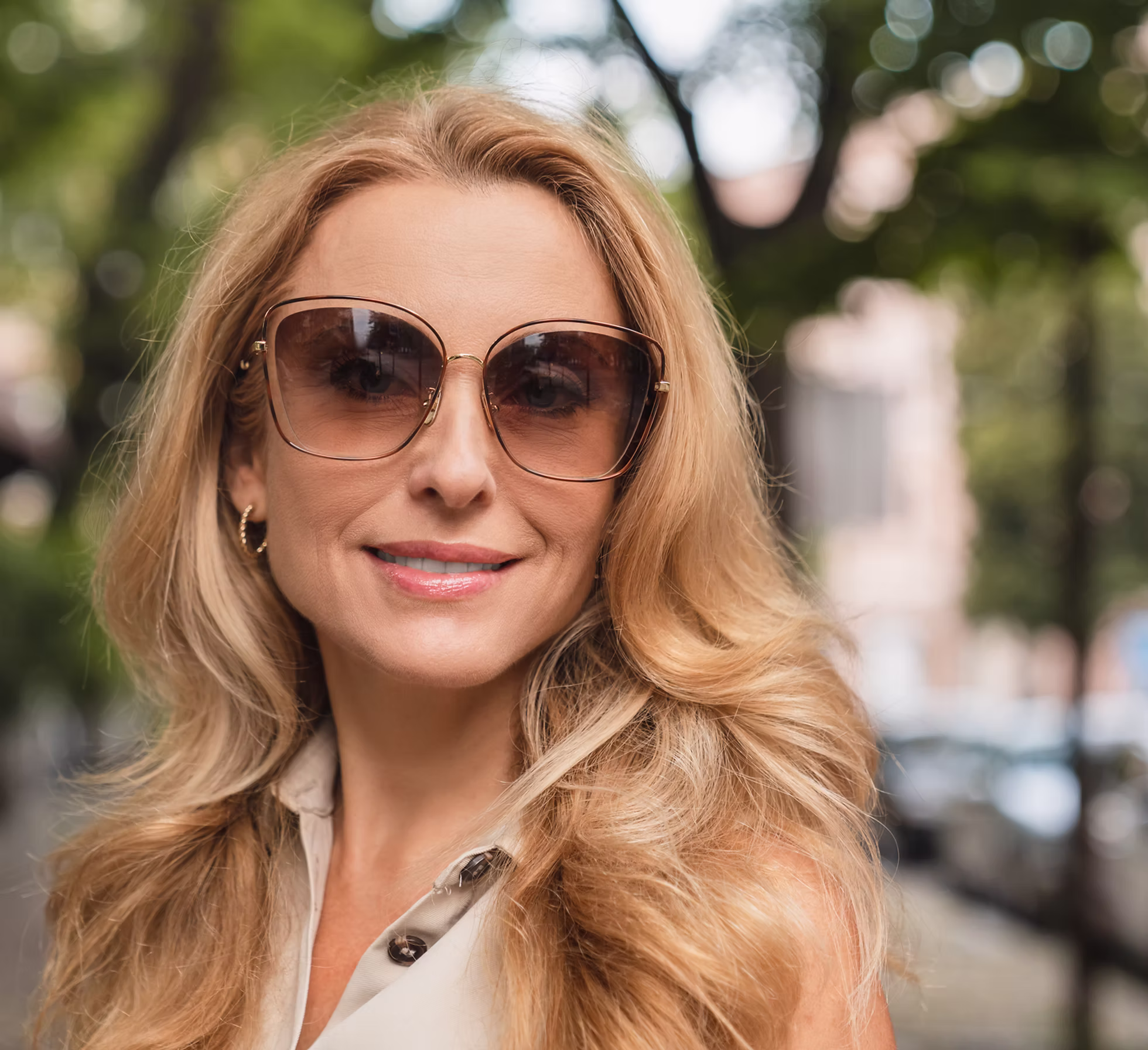 Smiling woman with long blonde hair wearing large brown sunglasses outdoors.