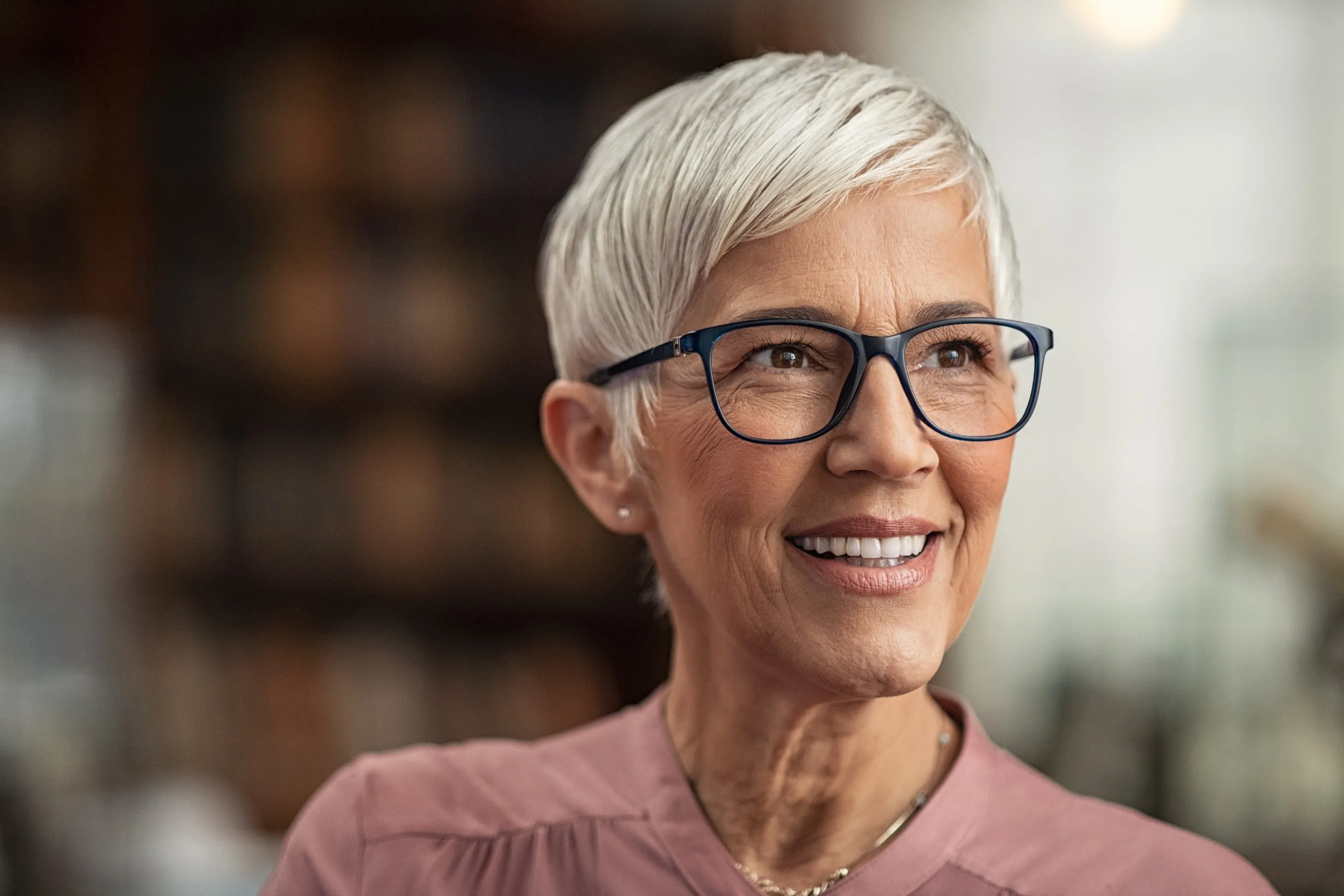 Smiling mature woman with short white hair and glasses wearing a pink top.
