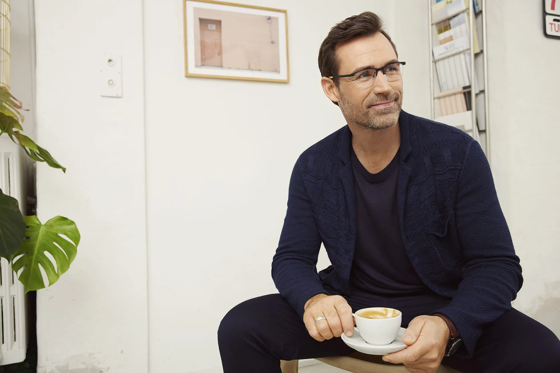 Smiling man wearing glasses and a navy cardigan holding a cup of coffee indoors.