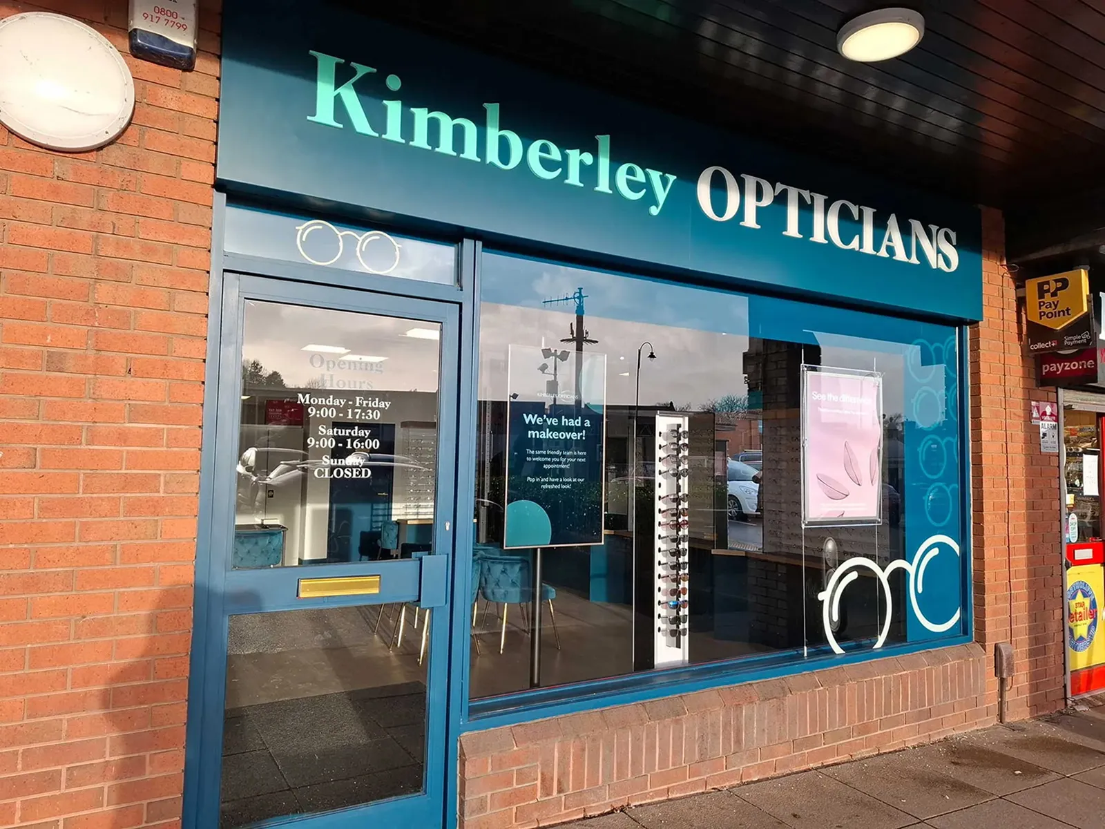 Exterior of Kimberley Opticians store with blue door, signage, and window displaying opening hours and promotional posters.