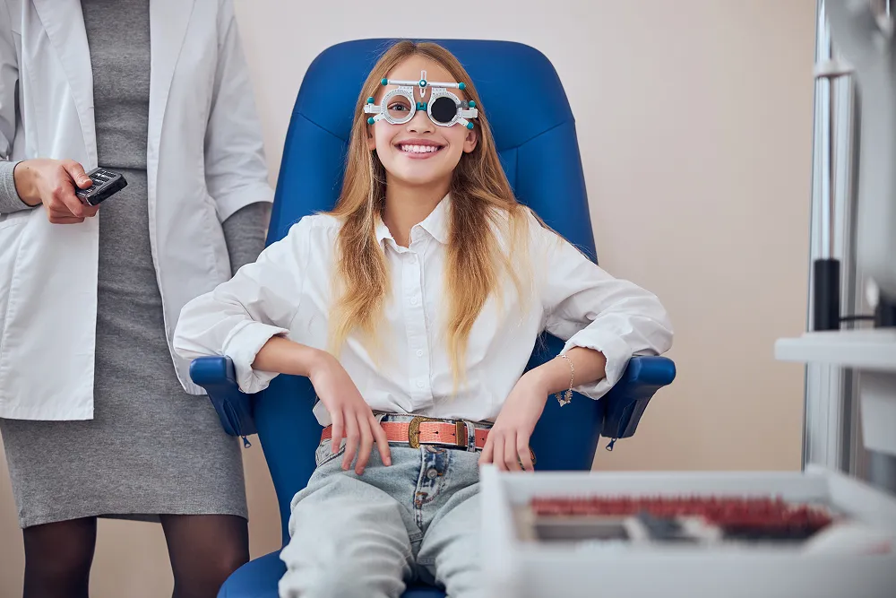 Smiling young girl wearing an eye testing device sitting in an optometrist's chair with a doctor standing beside her.