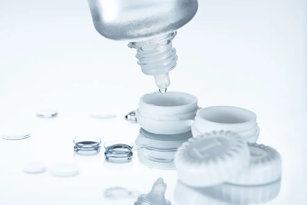 Contact lens case with a dropper releasing a liquid drop above it and contact lenses scattered nearby on a reflective surface.
