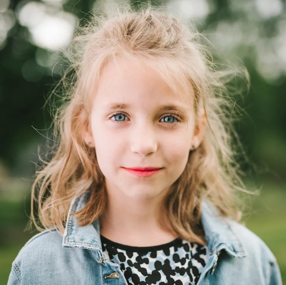 Close-up portrait of a young girl with blue eyes, light blonde hair, wearing a denim jacket and patterned shirt.