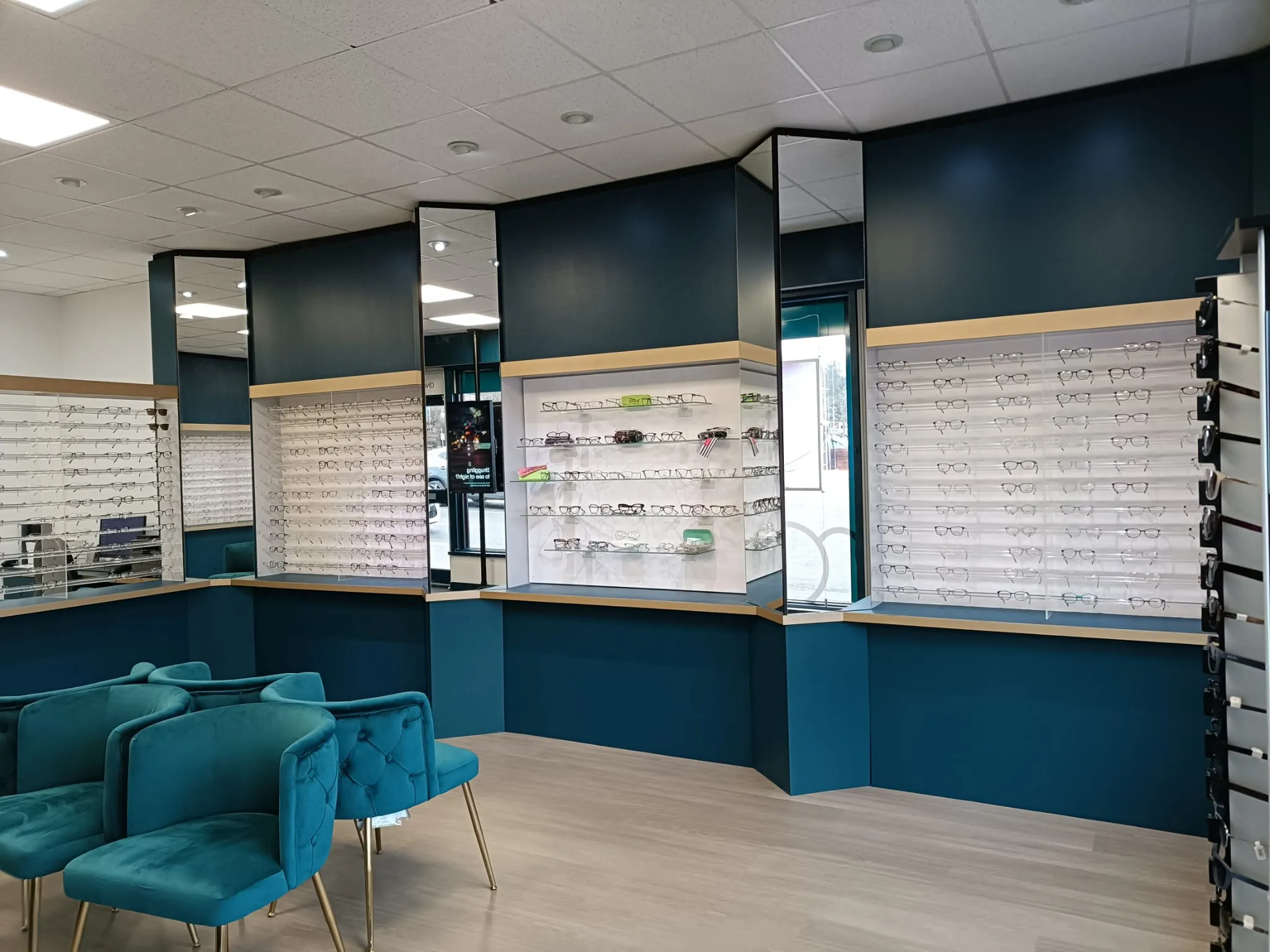 Interior of an eyeglass store with multiple eyeglass frames displayed on wall-mounted shelves and teal upholstered chairs in the seating area.