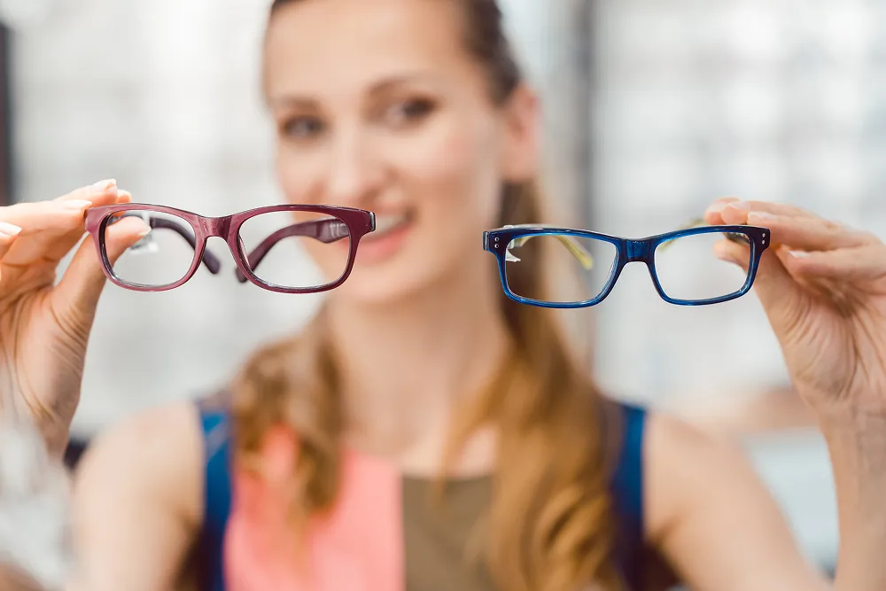 Woman holding a pair of burgundy eyeglasses in one hand and blue eyeglasses in the other, smiling.