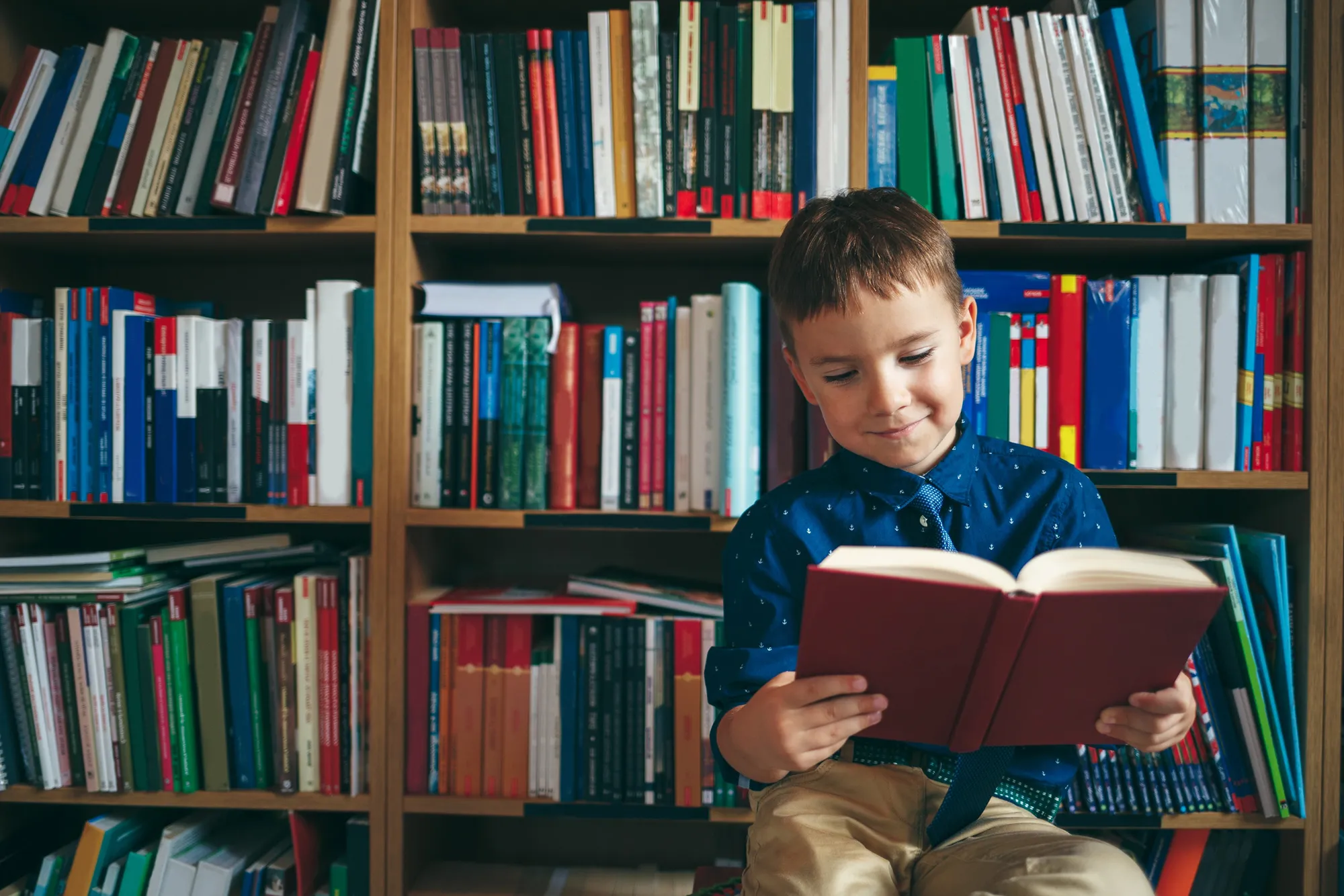 Young boy wearing a blue shirt and tie, sitting and reading a book in front of a filled wooden bookshelf.