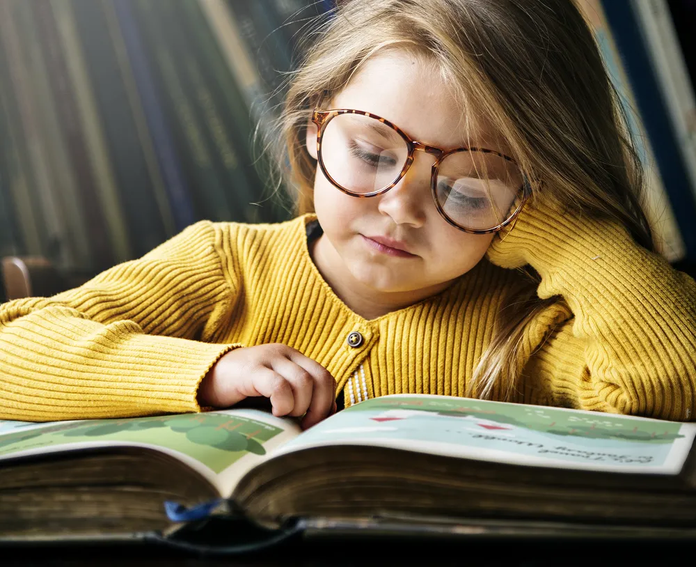 Young girl with glasses and a yellow sweater reading a large book.