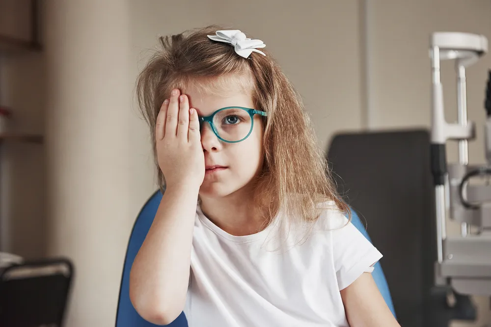 Young girl wearing glasses covers one eye with her hand during an eye test in an optometrist's office.