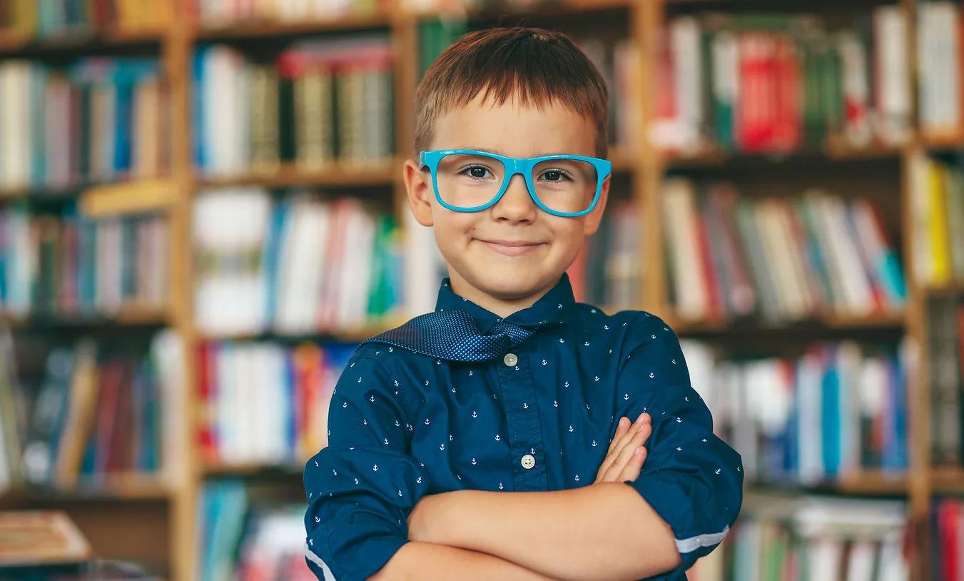 Smiling young boy wearing large blue glasses and a blue shirt with a bow tie in front of a bookshelf.