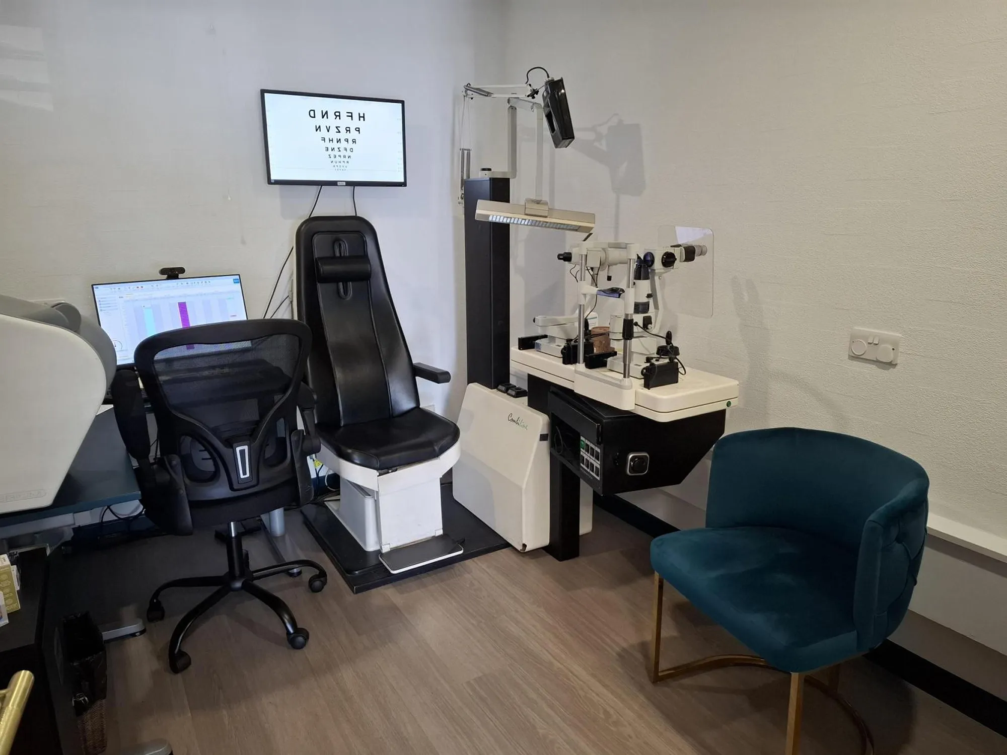 Modern optometry exam room with black exam chair, ophthalmic instrument, computer monitor, eye chart on wall, and a teal visitor chair.