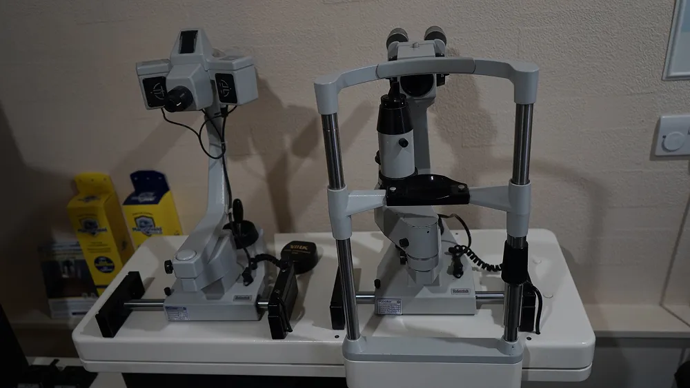 Two white ophthalmic devices with adjustable parts on a white table against a beige wall.