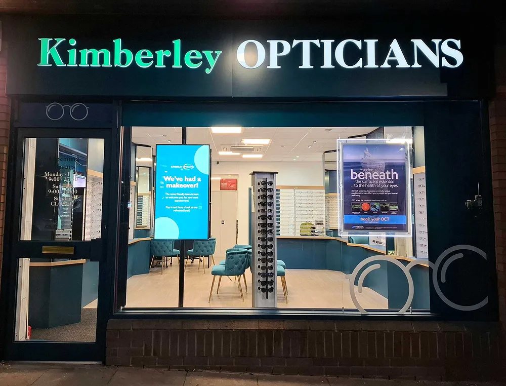 Front view of Kimberley Opticians storefront at night with illuminated sign and inside display of chairs, eyewear, and informational posters.