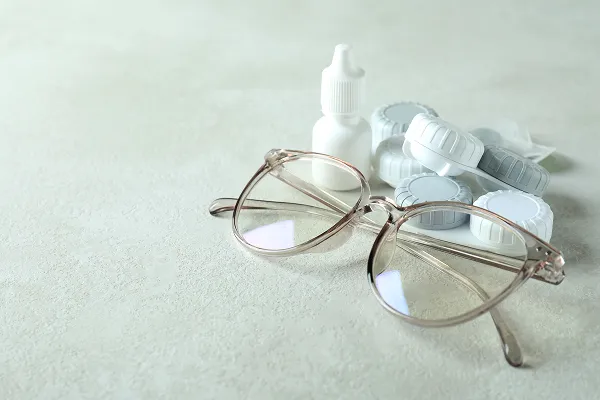 A pair of eyeglasses resting on a surface next to eye drop bottle and several white pills.
