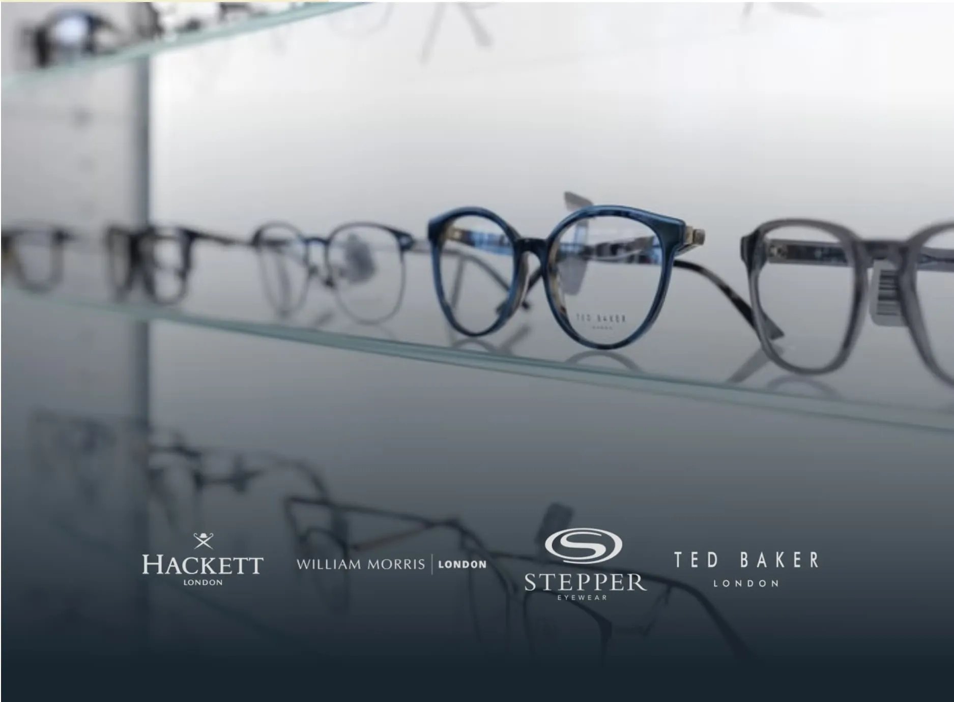A glass shelf display of designer eyeglass frames in various styles and shades of grey and blue, featuring brands Hackett London, William Morris London, Stepper Eyewear, and Ted Baker London