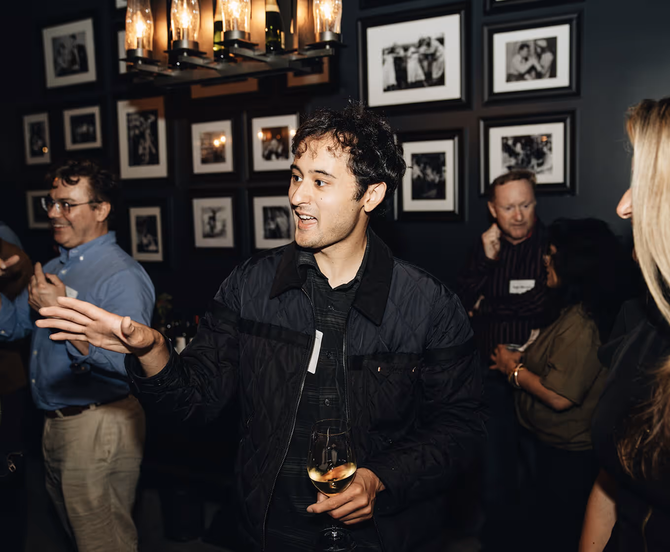 Gregg Mojica (CEO) holding a glass of white wine talking at a social gathering with people and framed photos in the background.