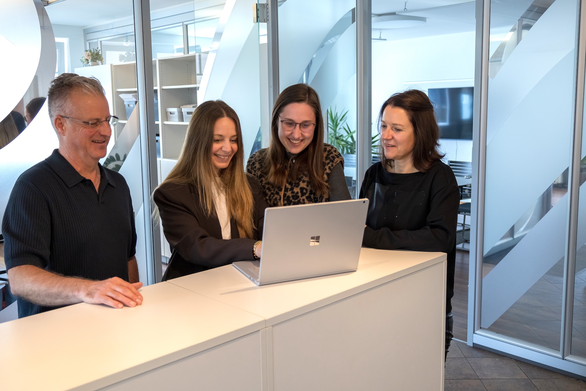 Vier lächelnde Personen schauen gemeinsam auf einen Laptop auf einem weißen Tisch im Büro.