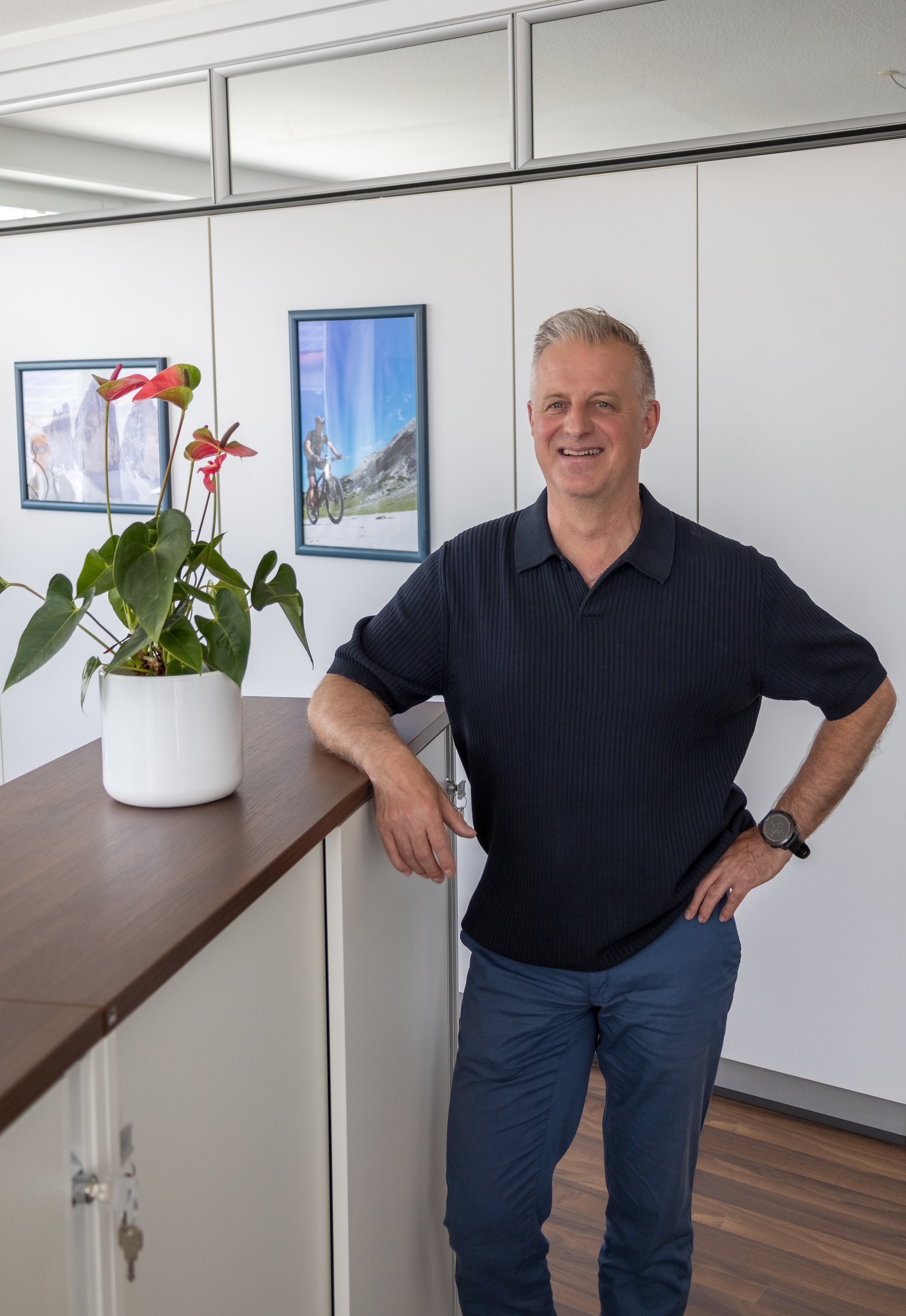 Ein lächelnder Mann lehnt an einem Schreibtisch im Büro neben einer Pflanze und Fotos an der Wand.