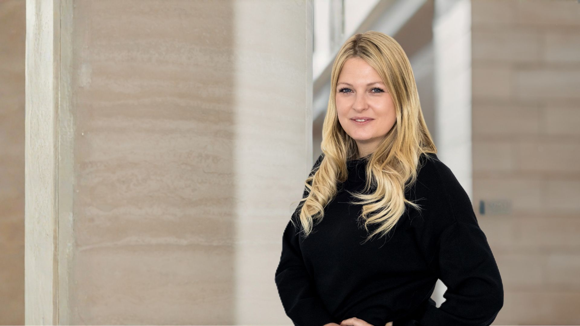 Blonde Frau mit lockigen Haaren trägt schwarzen Pullover und steht vor hellem Mauerhintergrund.
