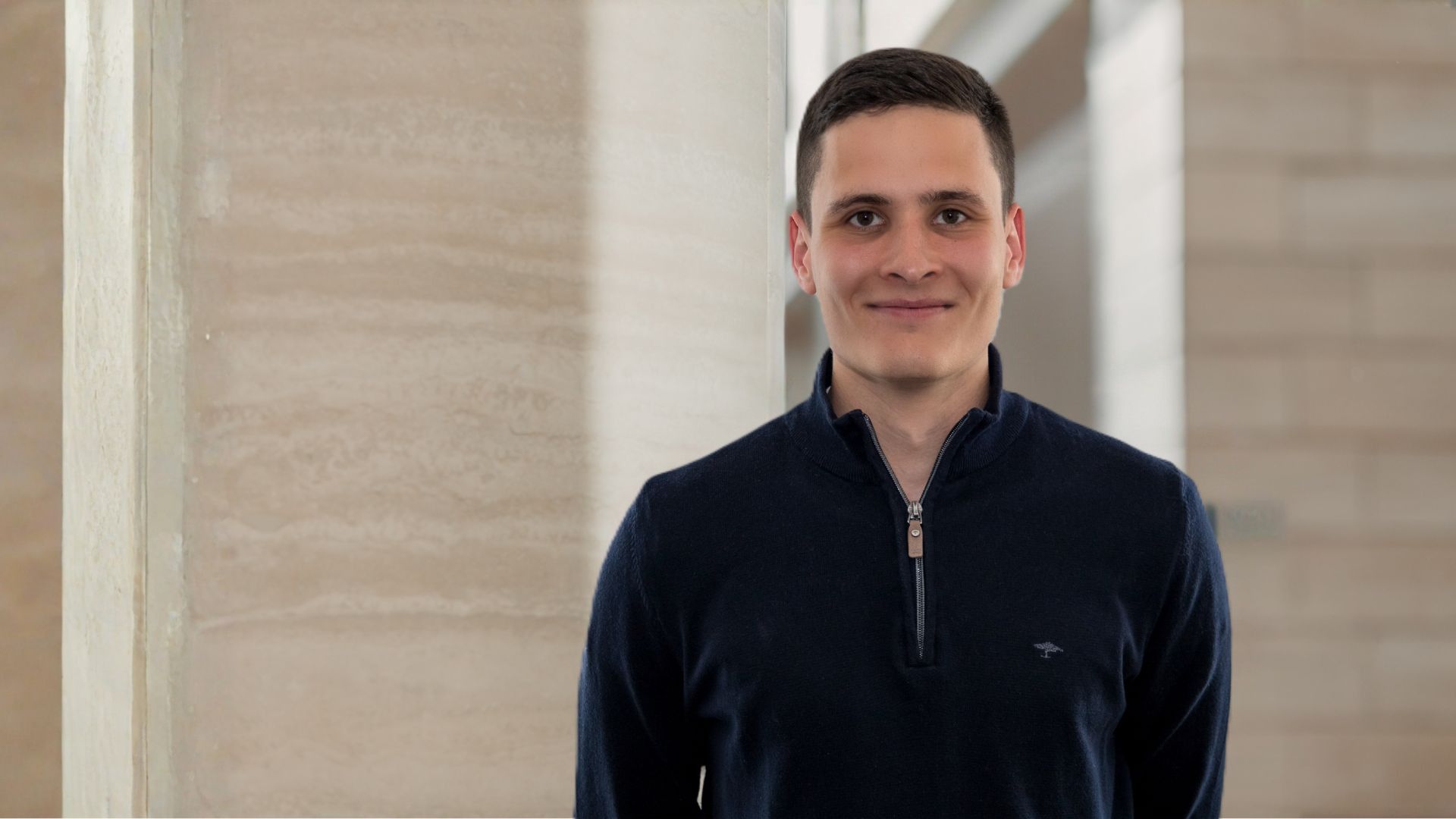 Junger Mann mit kurzem dunklem Haar lächelt vor einem beige-grauen Hintergrund aus Steinwänden.