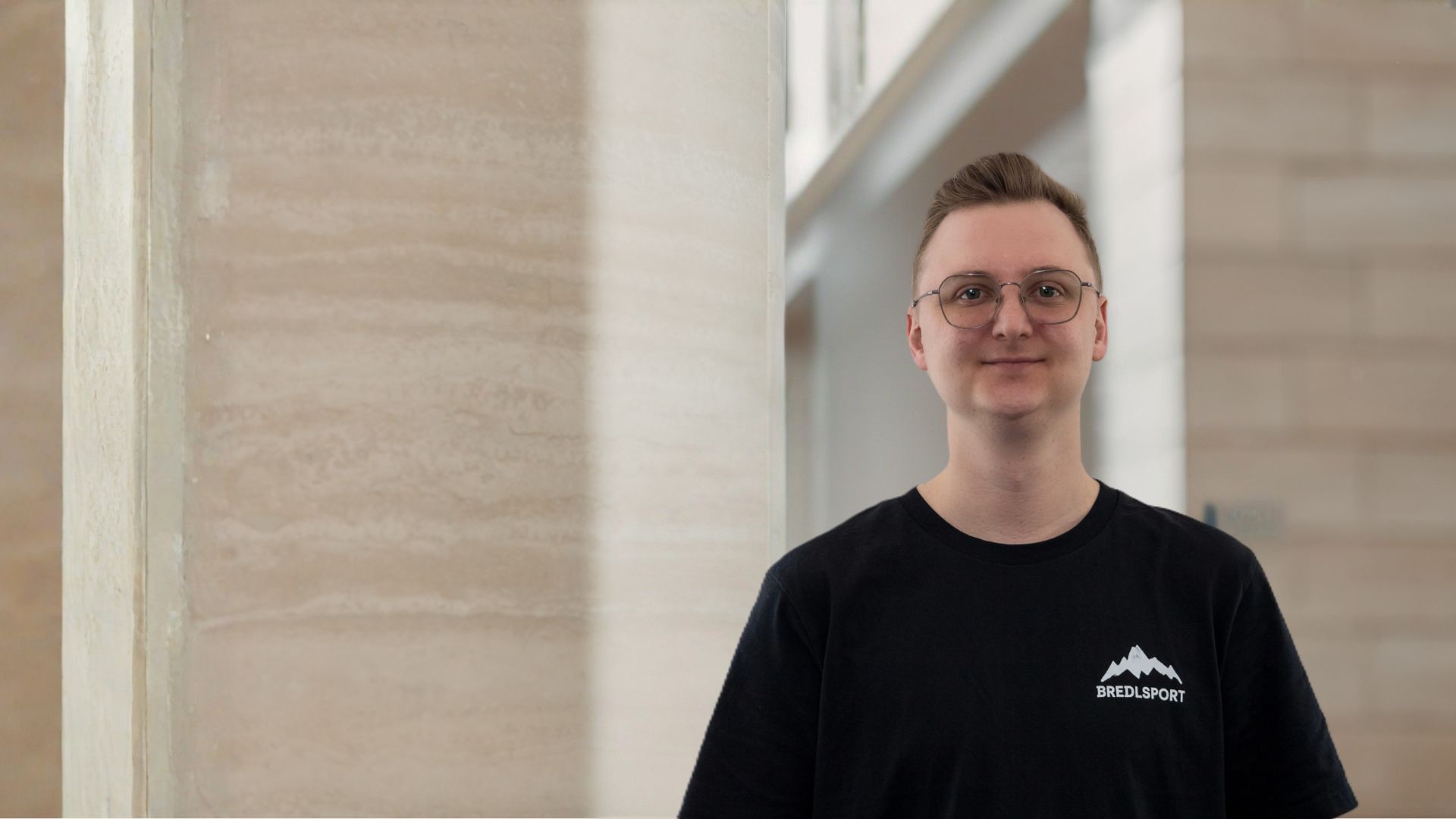 Junger Mann mit Brille trägt schwarzes T-Shirt mit Bredlsport-Logo vor heller Wand.