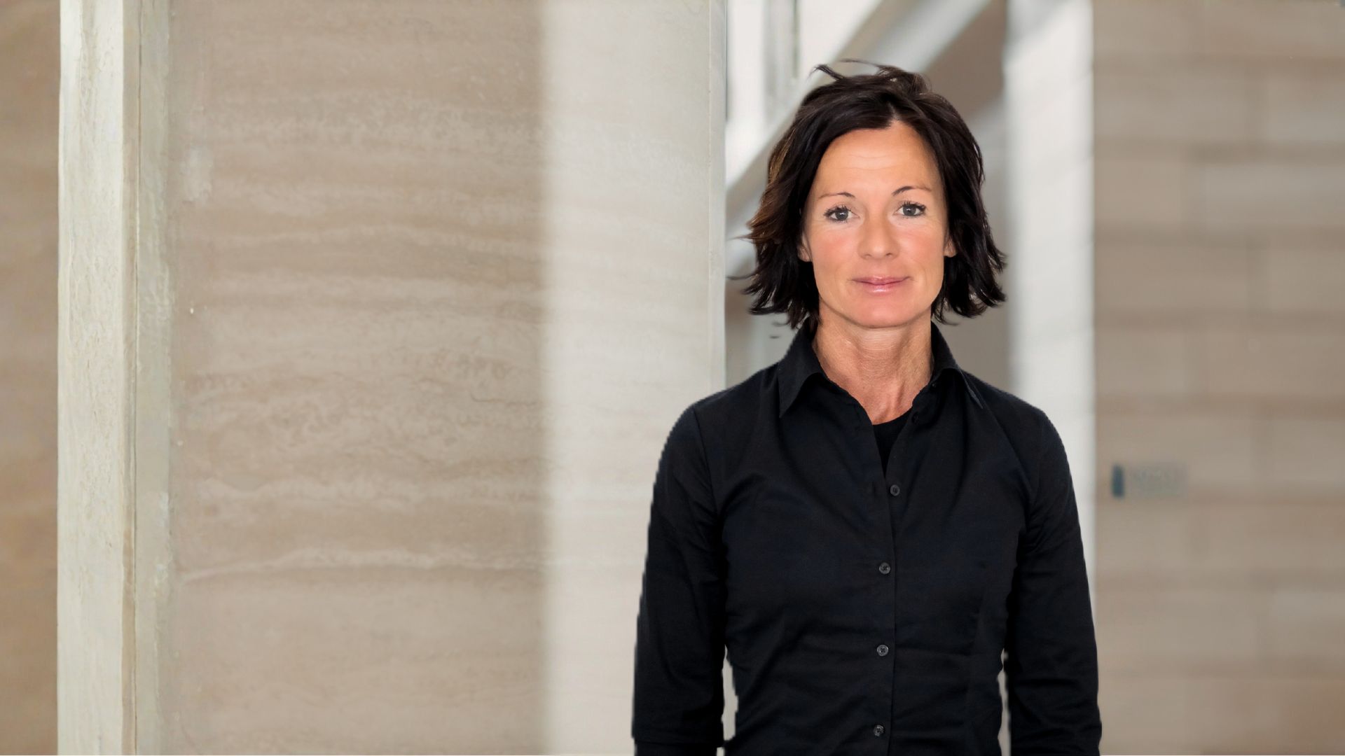 Frau mit kurzen braunen Haaren und schwarzem Hemd vor hellem Steinwandhintergrund.