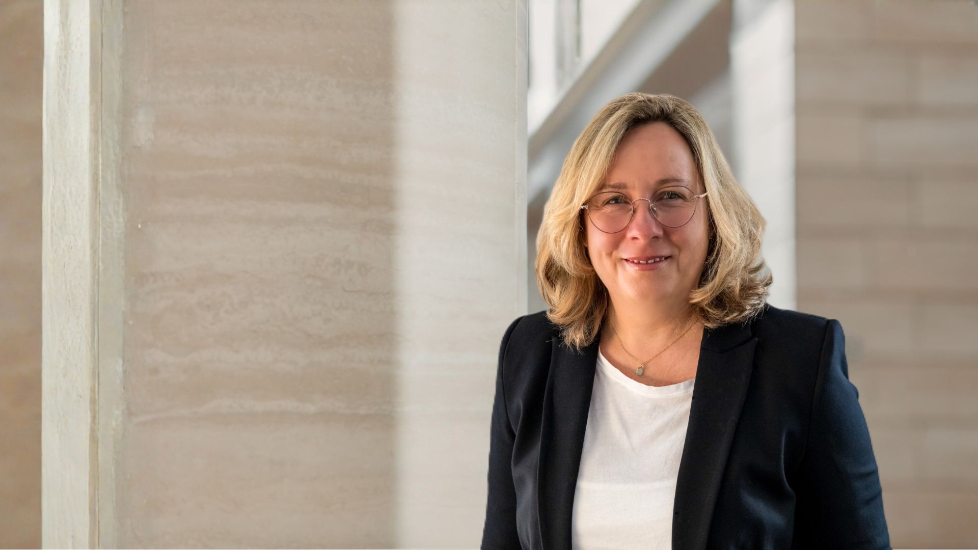 Lächelnde Frau mit blonden Haaren, Brille, weißem Shirt und dunklem Blazer vor heller Wand.