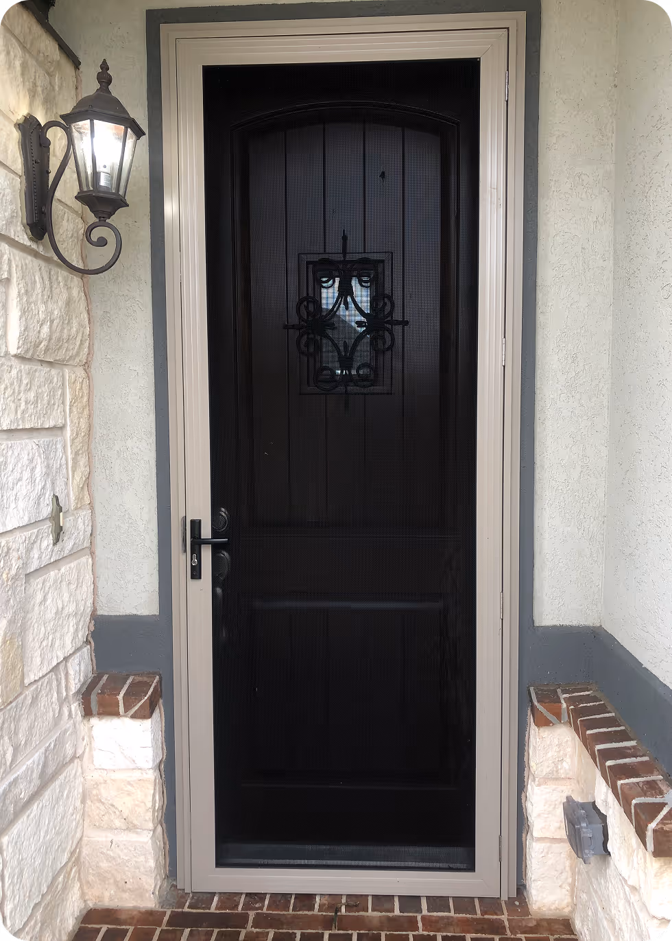 Black front door with decorative wrought iron grill in a beige frame, flanked by stone walls and a wall-mounted lantern light on the left.