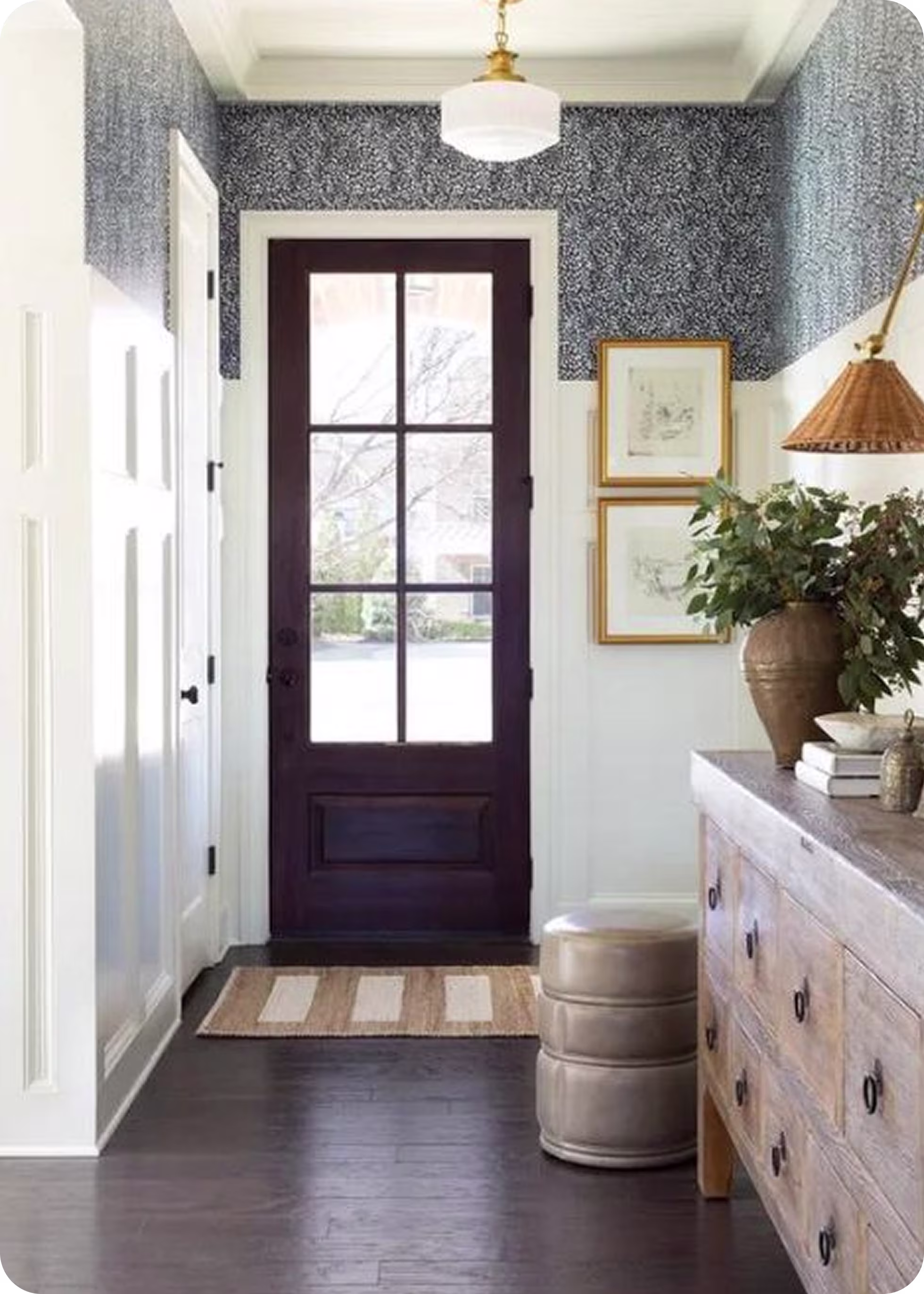 Entryway with a dark wood door featuring glass panels, striped area rug, light-patterned wallpaper above white wainscoting, wooden dresser, and a round beige ottoman.