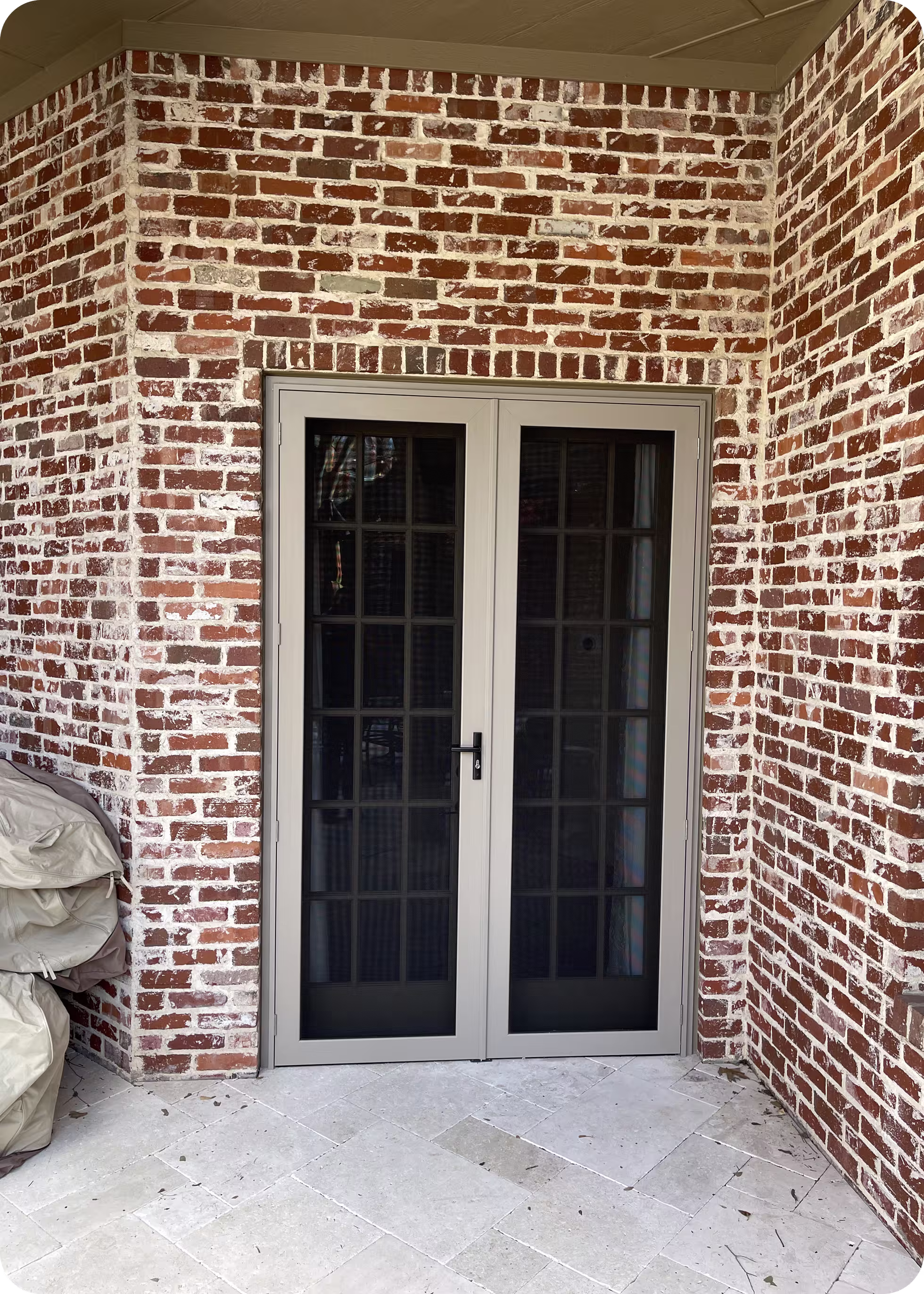 Closed double glass doors with white frames set in a red brick wall on a light stone tile patio.