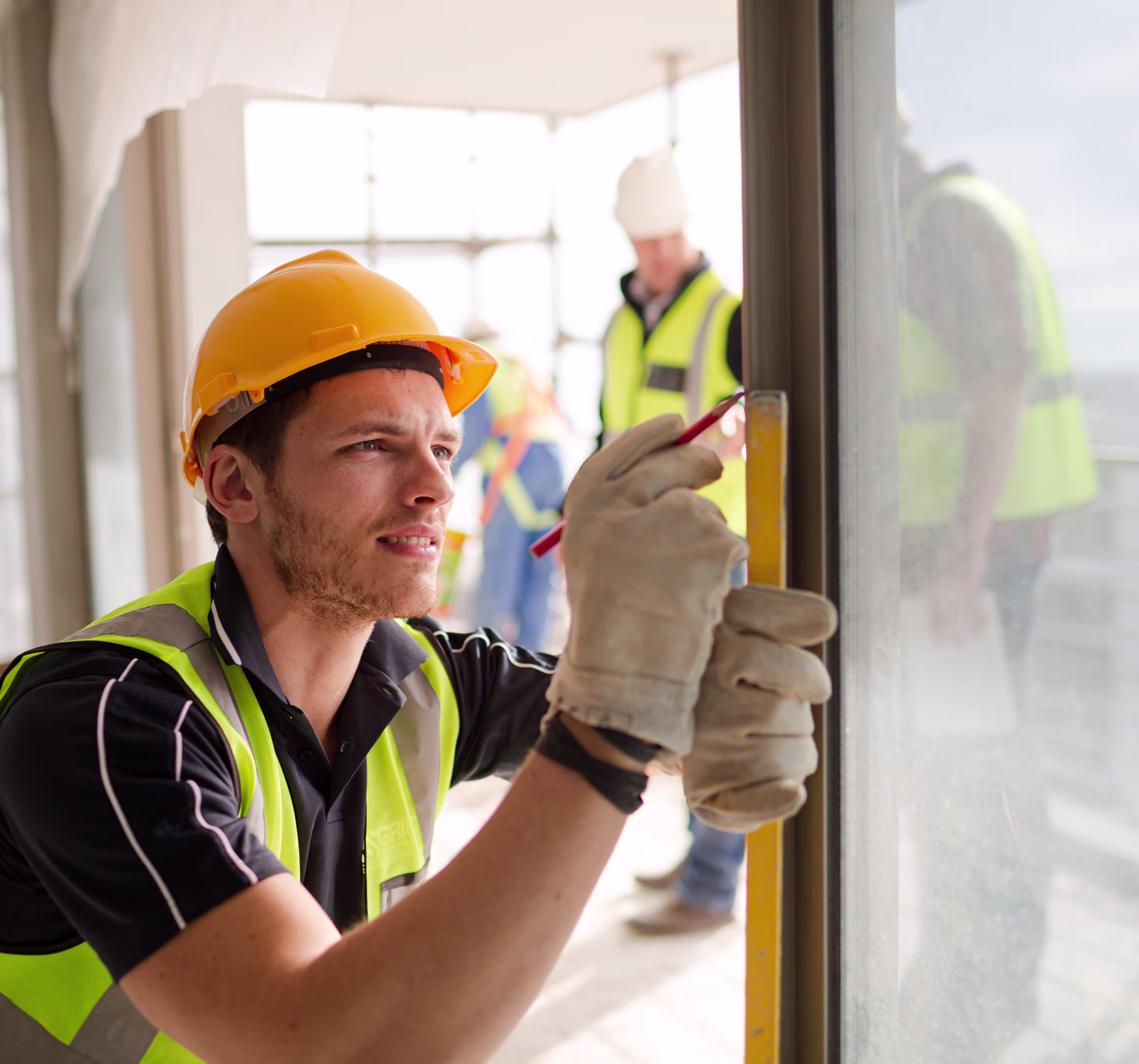 Construction worker in a yellow hard hat and safety vest marking a window frame with a pencil while holding a level.