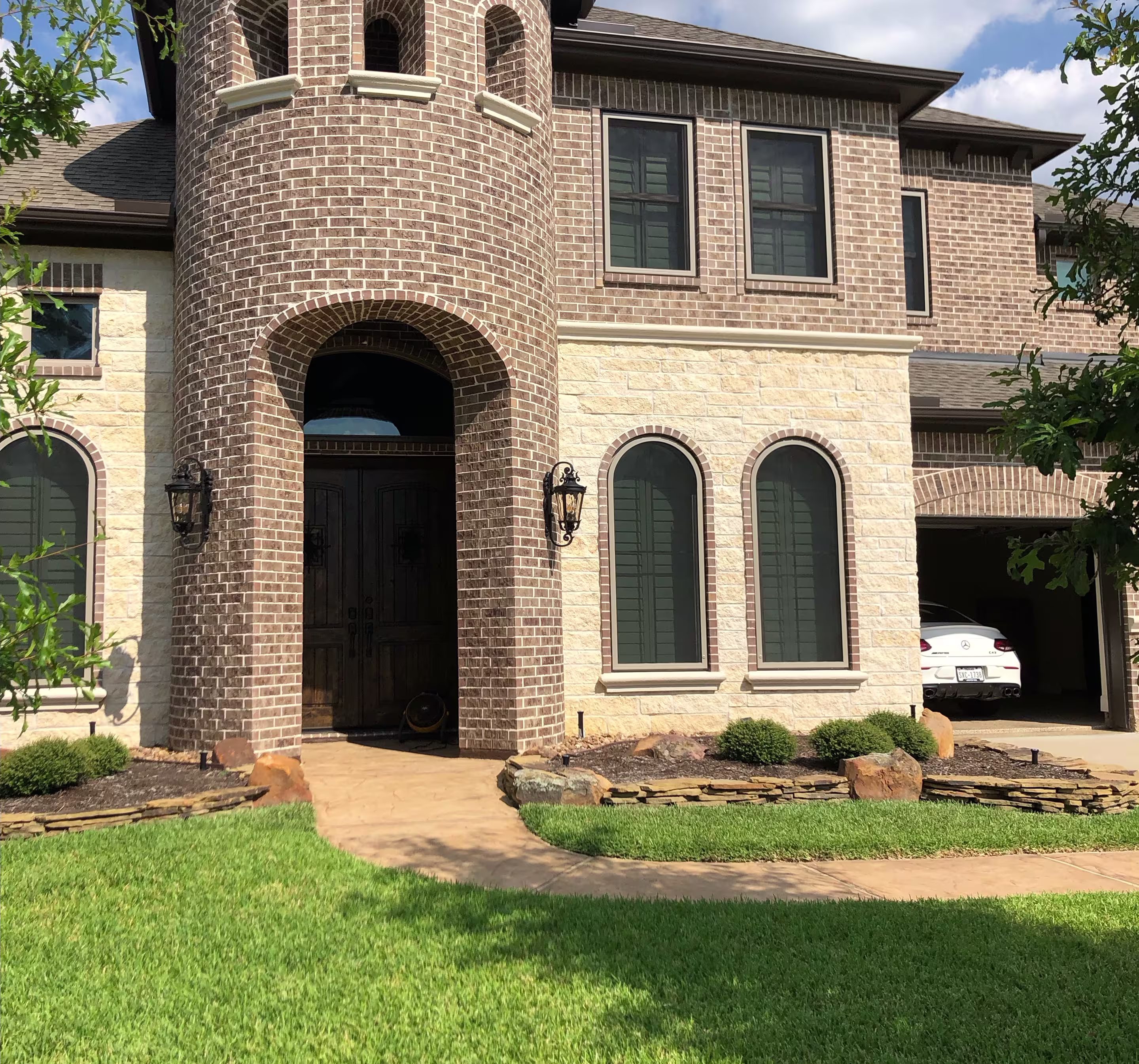 Two-story brick and stone house with arched windows, a rounded brick entryway, and a driveway leading to a garage with a white car.