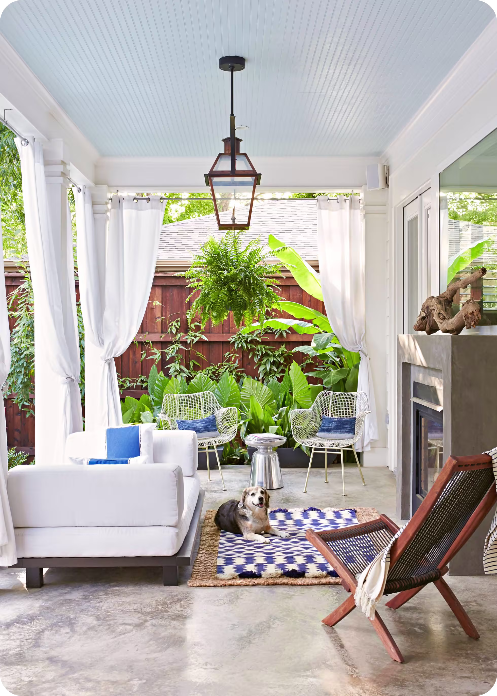 Bright covered patio with white sofas, two wire chairs, a hanging fern, blue and white rug, and a dog lying on the rug.
