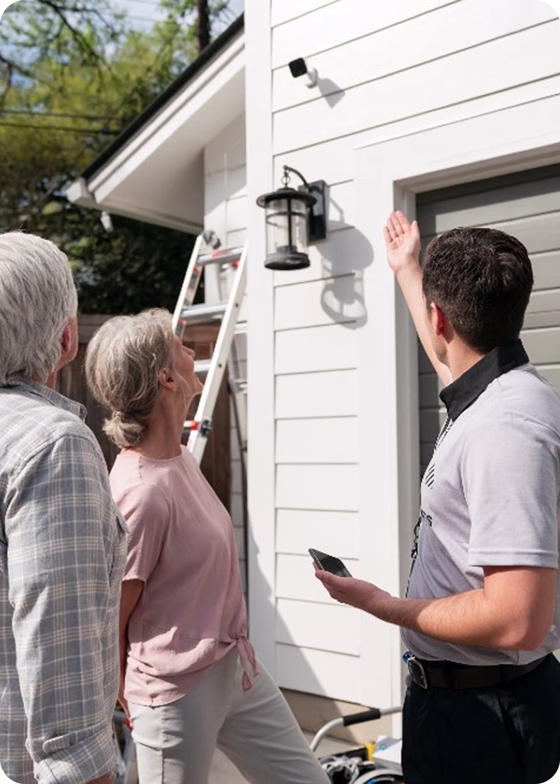 A man showing an older couple a security camera and outdoor lantern on a house exterior.