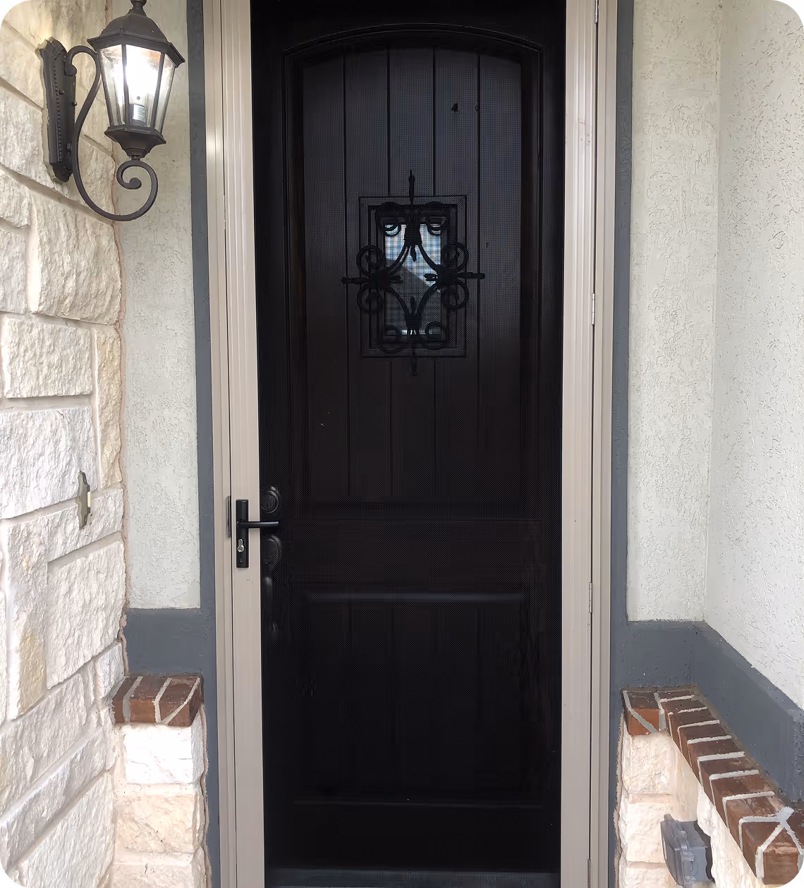 Black front door with decorative metal grille in a beige frame, flanked by light stone and brickwork, with a lit wall lantern on the left.