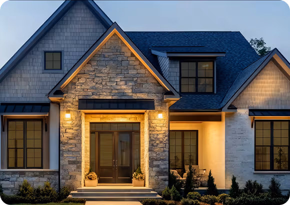Modern house exterior at dusk with stone and siding facade, illuminated entryway, and large windows.