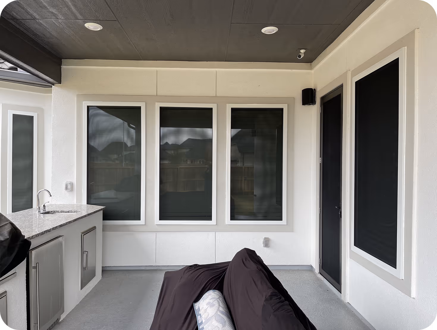 Covered patio with three large windows, a door, a countertop with a sink, and a covered sofa facing the windows.