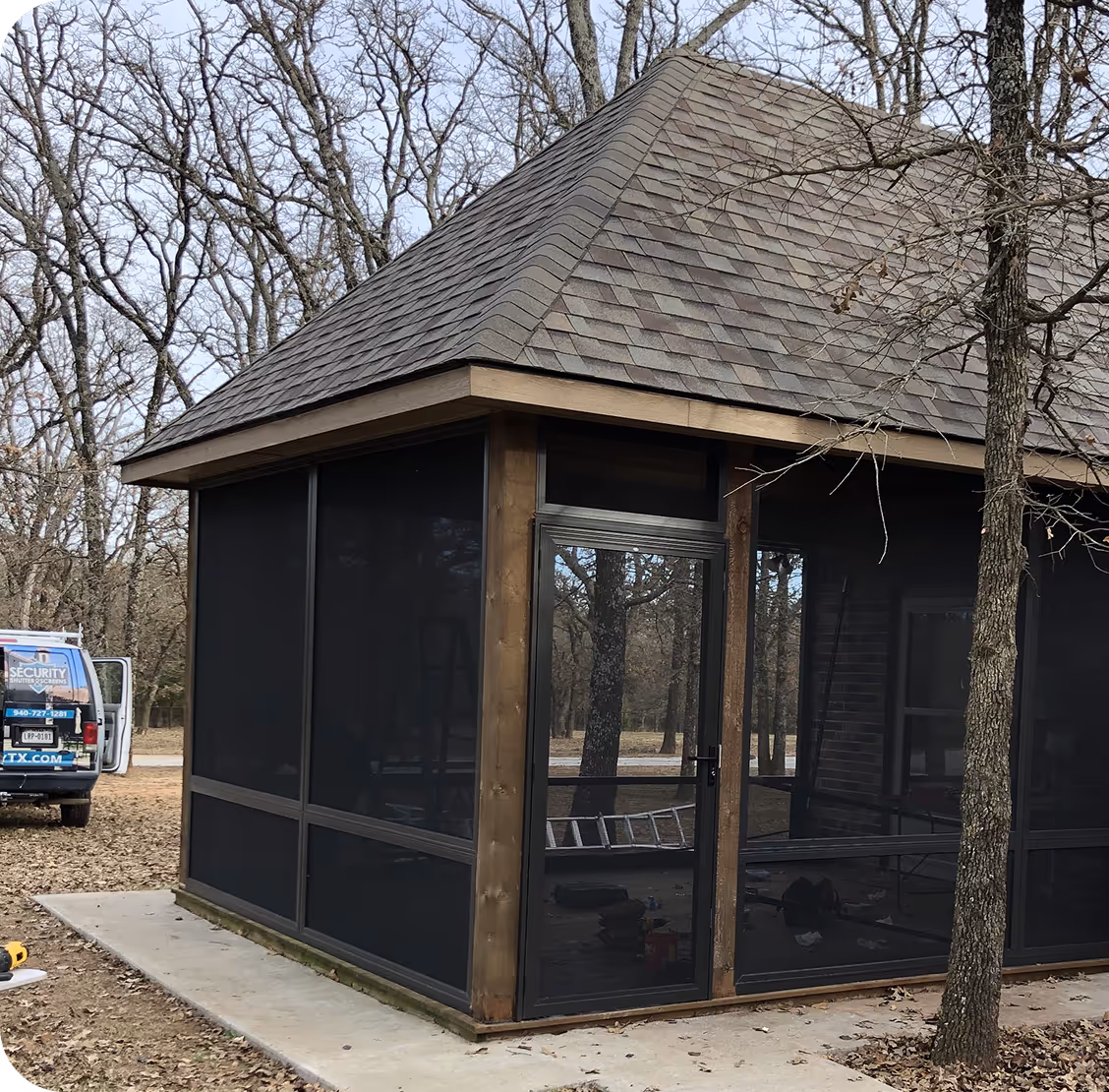 Wood-framed screened patio with a brown shingled roof on a concrete slab surrounded by leafless trees.