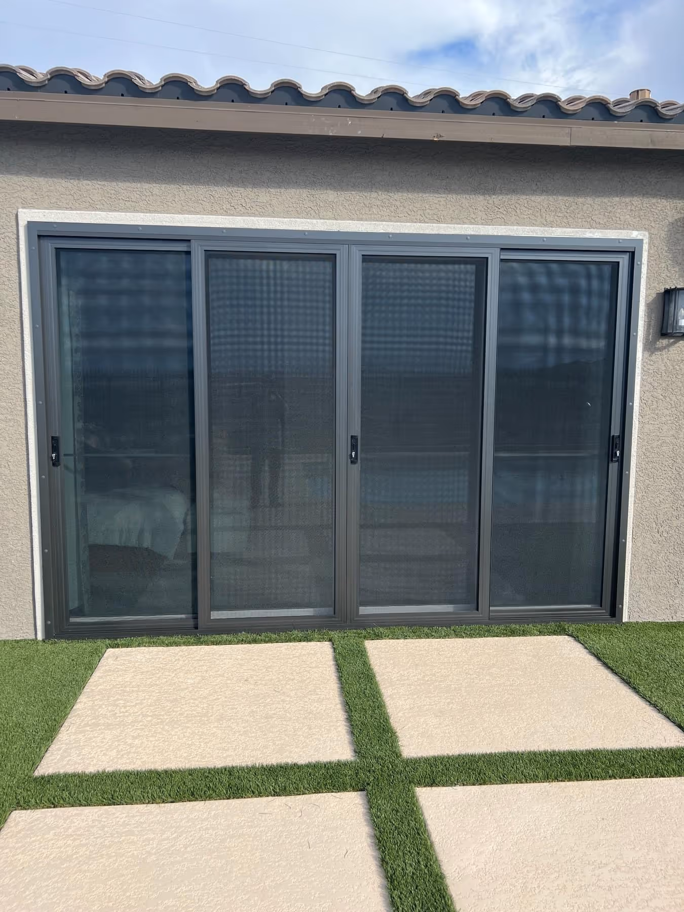 Four-panel dark gray sliding glass door with screen doors on a beige stucco wall above a patio with square concrete slabs and artificial grass.