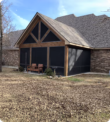 A wooden-framed screened porch attached to a brick house with a bench and some shrubs in front.