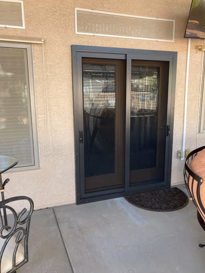Sliding glass door with black frame on a patio, with a patterned doormat in front and part of a wrought iron chair visible on the right.