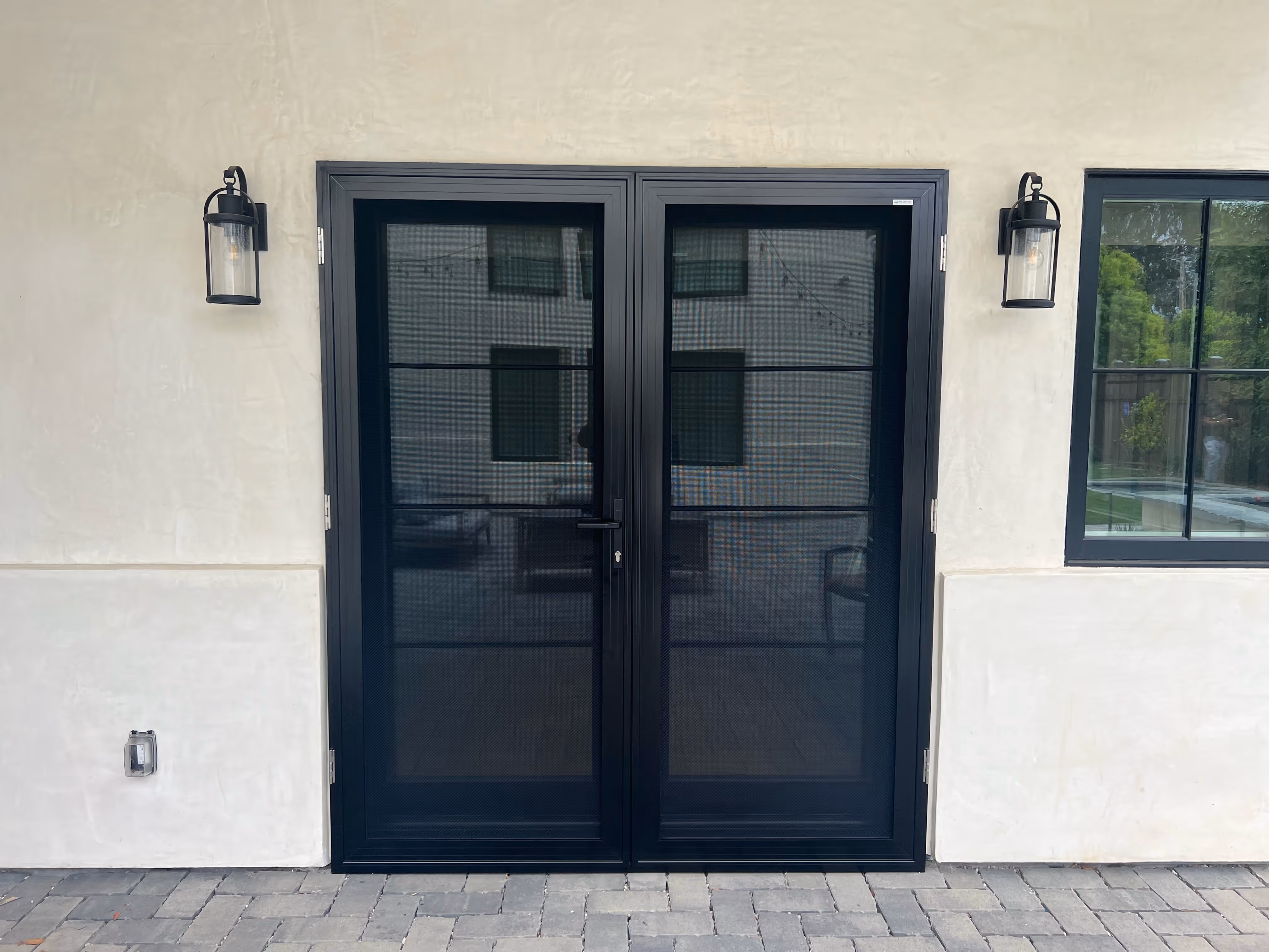 Modern black-framed double glass door with mesh screens, mounted on a light beige stucco wall between two black lantern-style wall lights and next to a window.